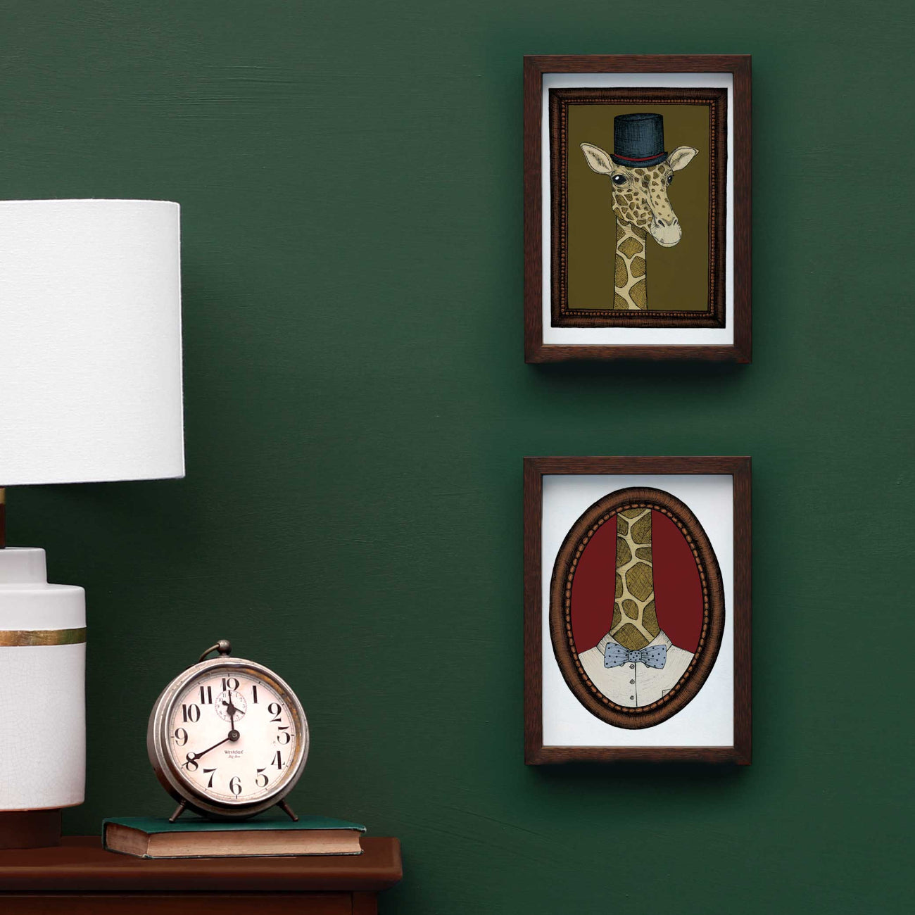Two framed artworks of giraffes on a green wall with a lamp and clock below.