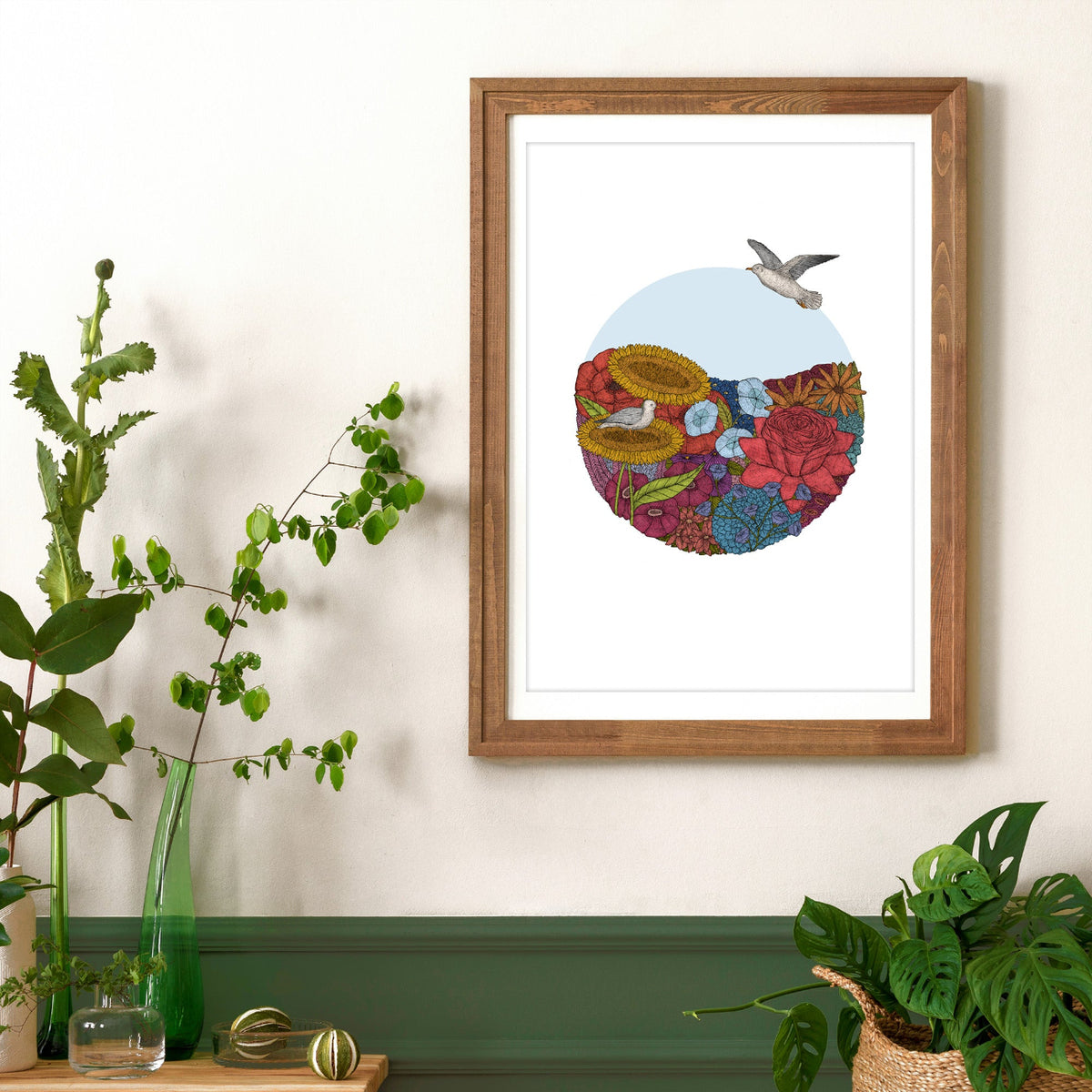 Framed artwork with a colorful design of flowers and birds on a wall with plants below.