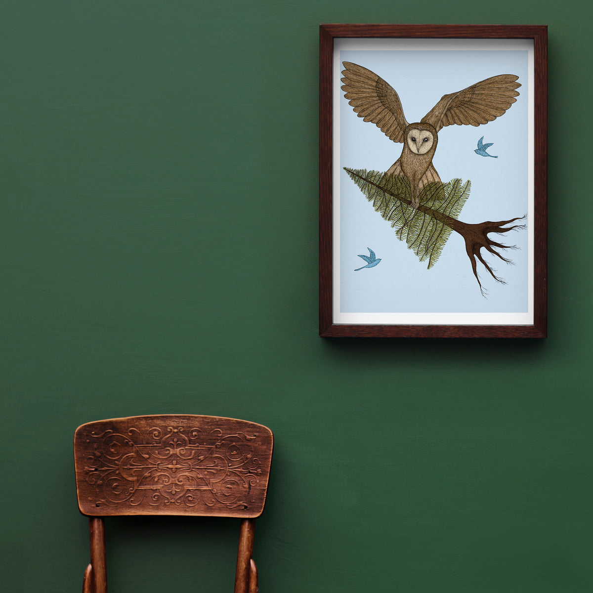 Framed artwork of an owl on a branch against a green wall with a wooden chair below.