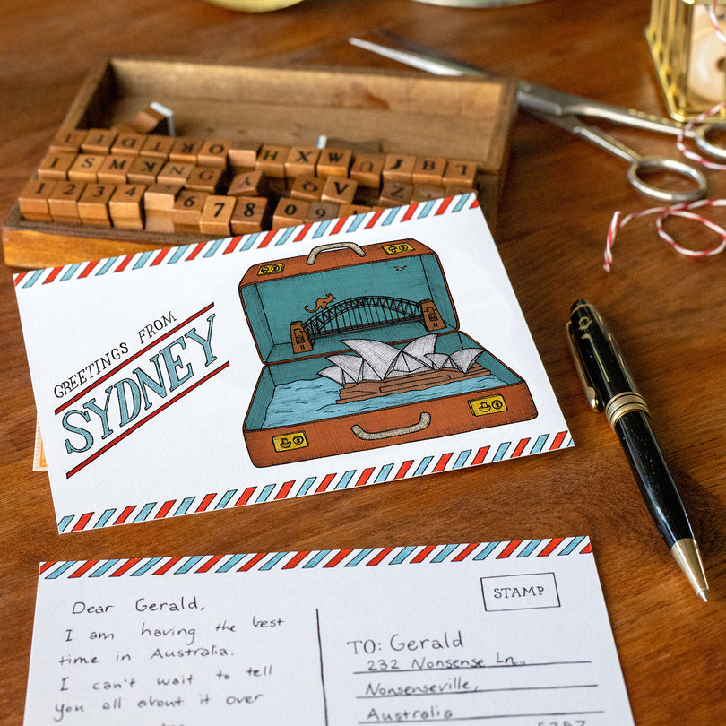 Postcard with an illustration of a suitcase containing the Sydney Opera house and Harbour Bridge, surrounded by stationery items on a wooden surface.