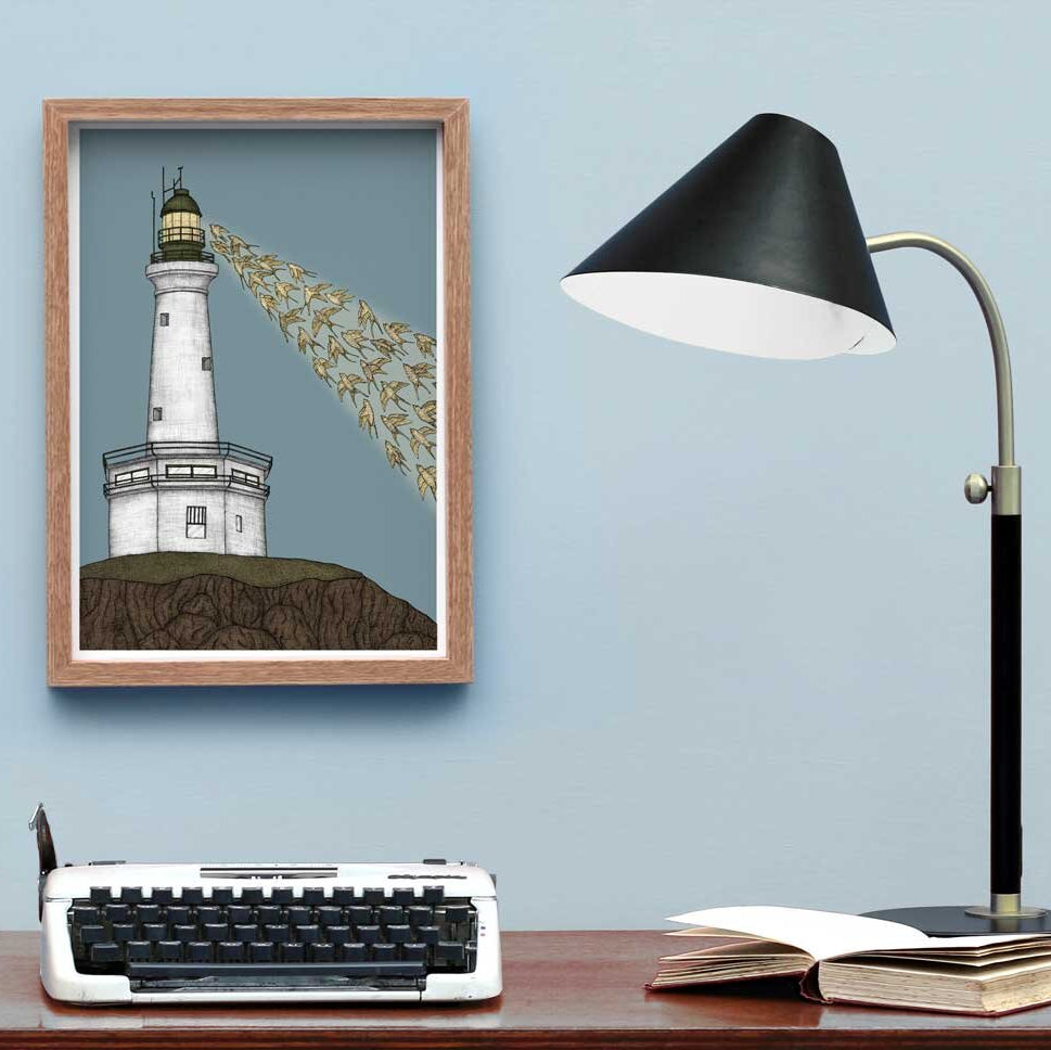 An illustration of a lighthouse with birds, framed and displayed on a wall next to a desk with a lamp and typewriter.