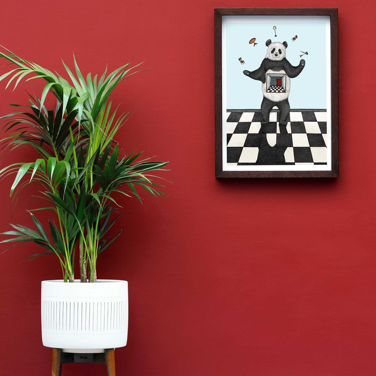 Limited edition art print featuring a whimsical illustration of a panda juggling objects, framed, with a red wall in the background and a white plant pot on the side.