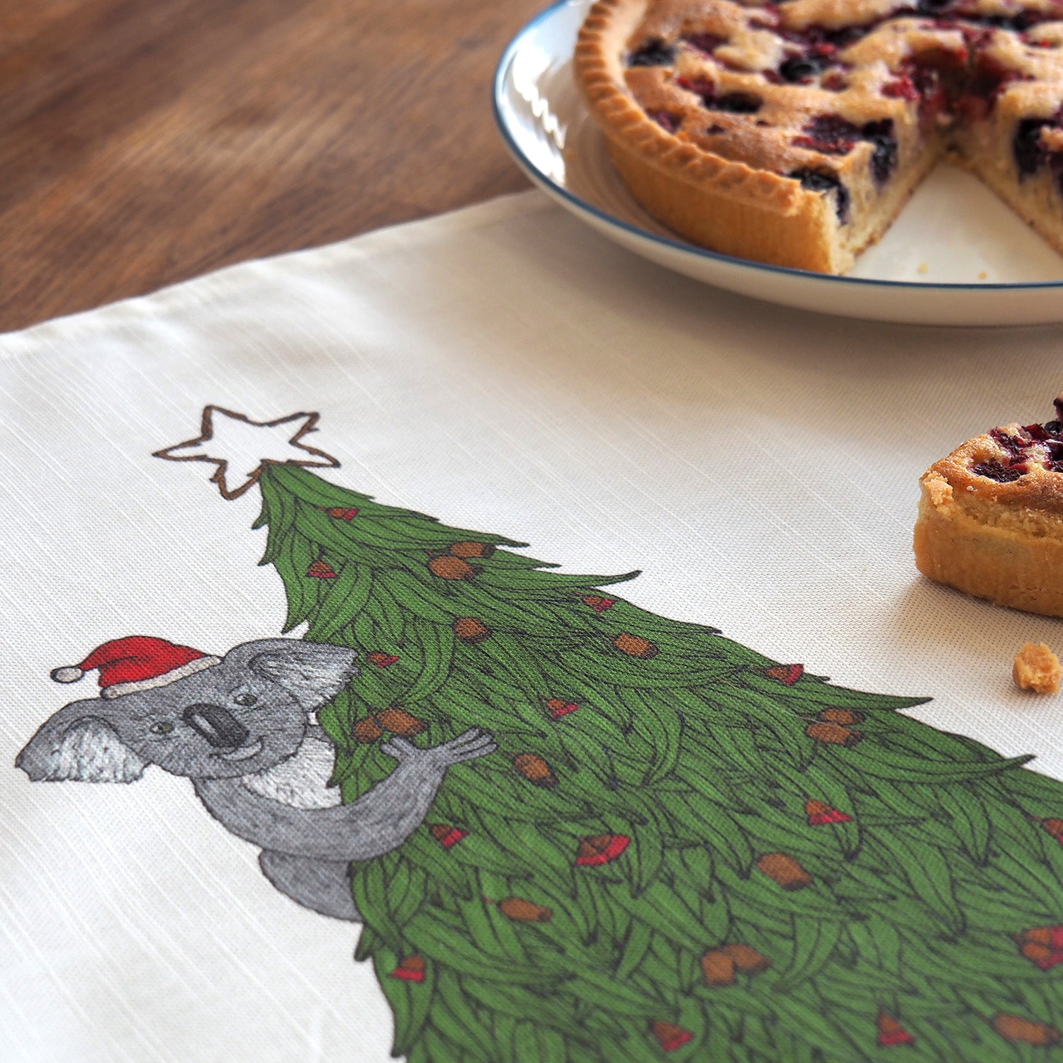 Tea towel with a koala in a koala and aChristmas tree design on a table with a pie.
