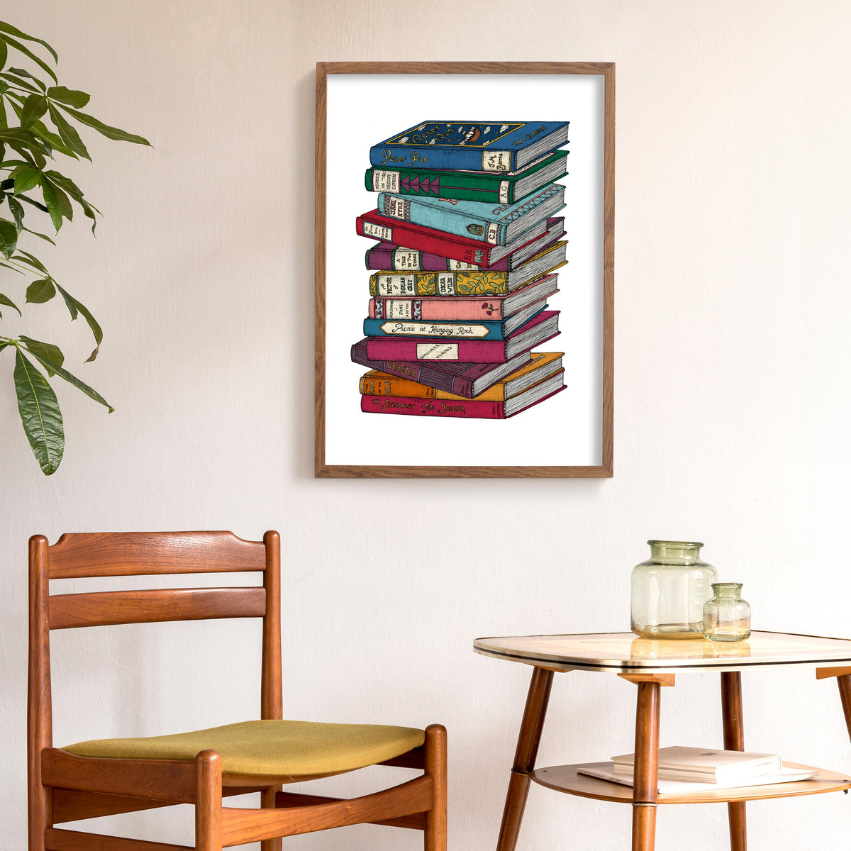 Framed artwork of a stack of colorful books on a wall above a wooden chair and table.