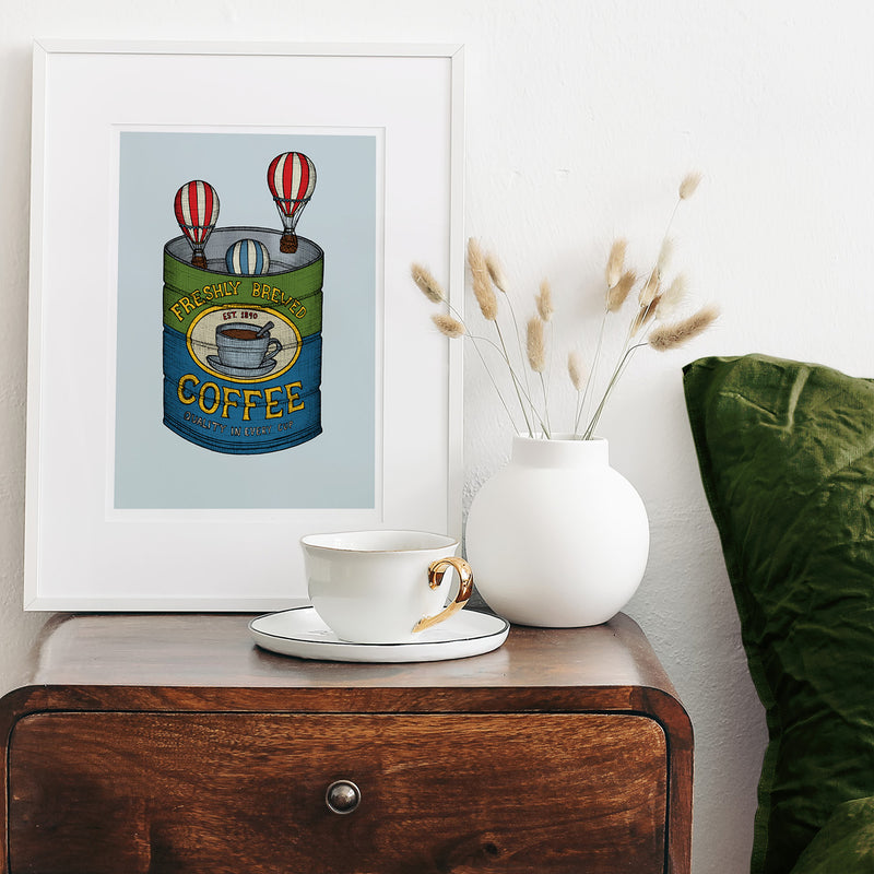 Framed artwork of a coffee can with hot air balloons, placed on a wooden surface with a cup and vase.