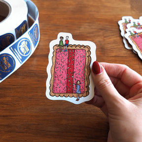 Hand holding a magnet with a design of people on an iced vovo, with stickers in the background.