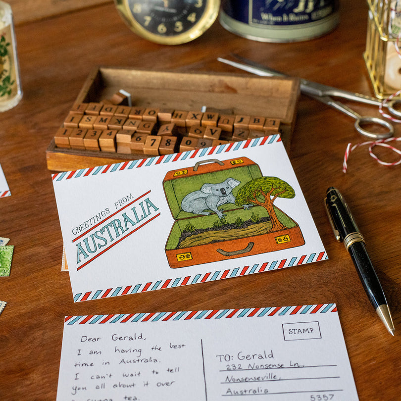 Postcard with a koala illustration and 'Greetings from Australia' text on a wooden surface with stationery items.