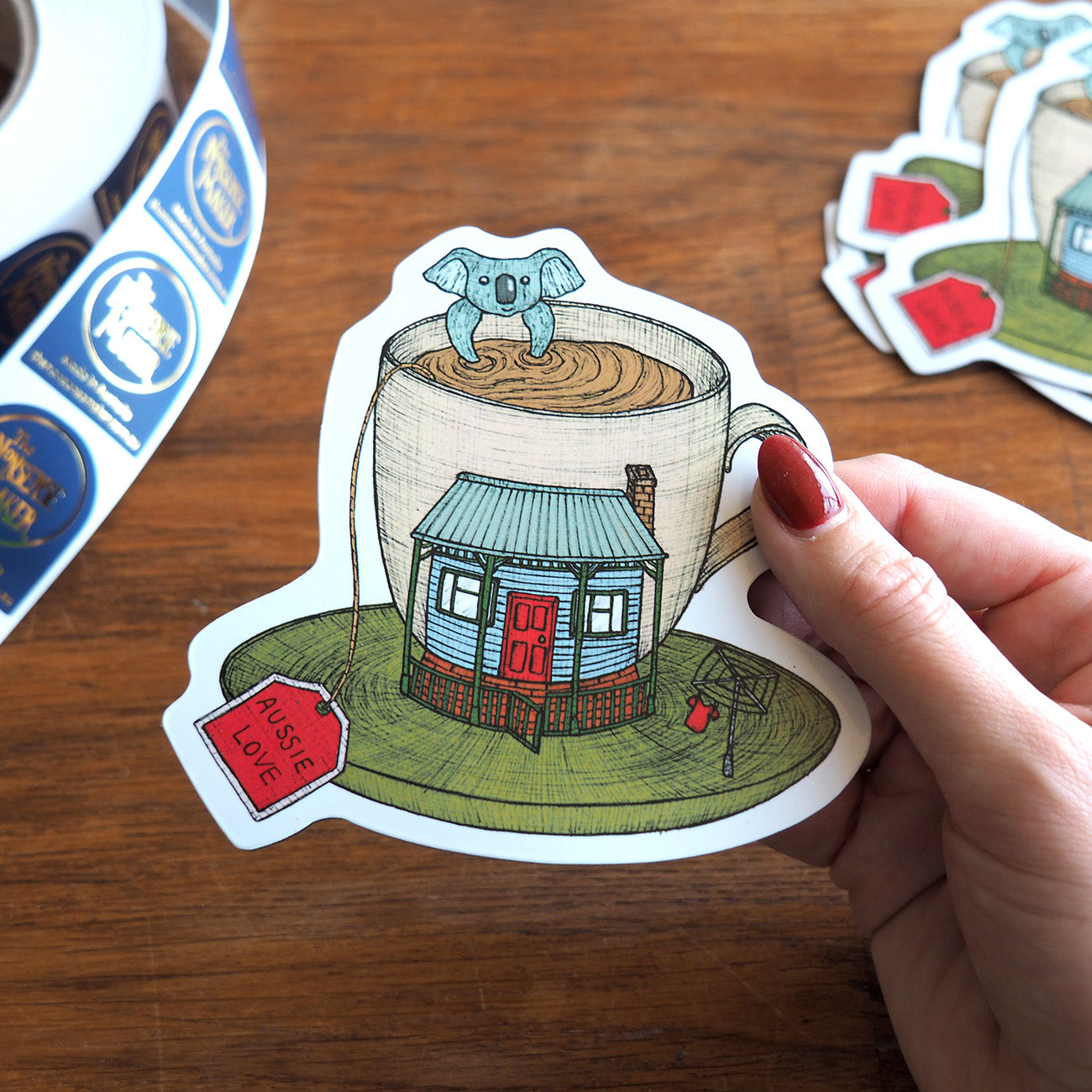 Magnet of a house in a coffee cup with a koala, held by a hand on a wooden surface.