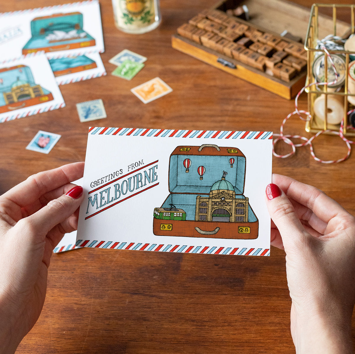 Postcard with 'Greetings from Melbourne' design held by hands on a wooden surface with stationery items.