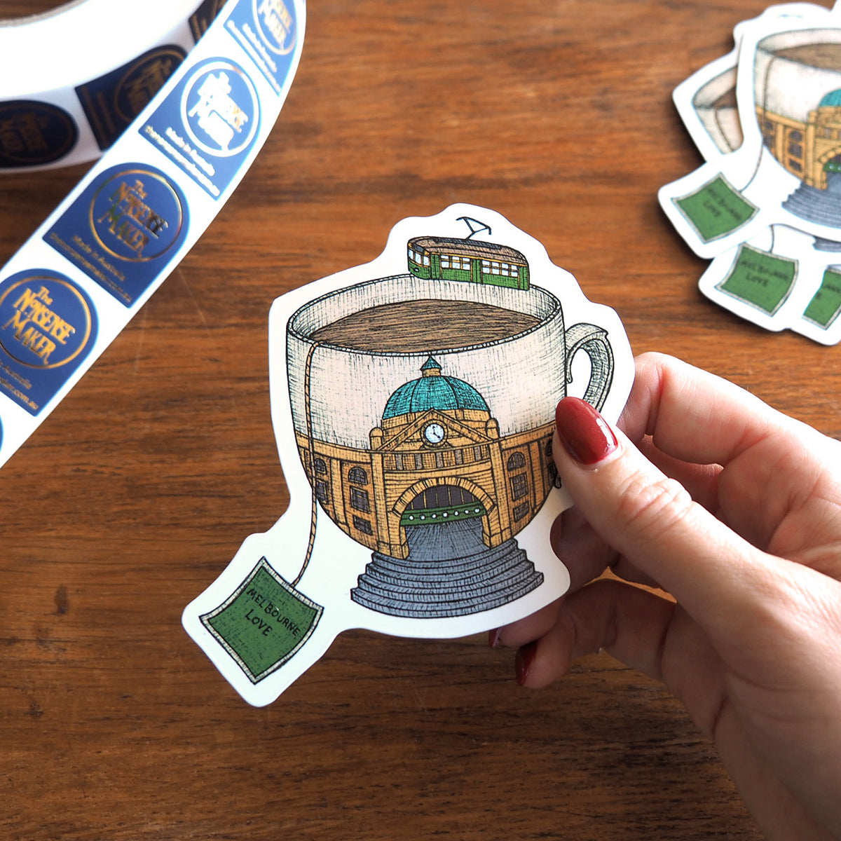 Magnet with an illustration of a Flinders street station inside a large tea cup with a tea bag labelled Melbourne love. Being packaged on a wooden table.