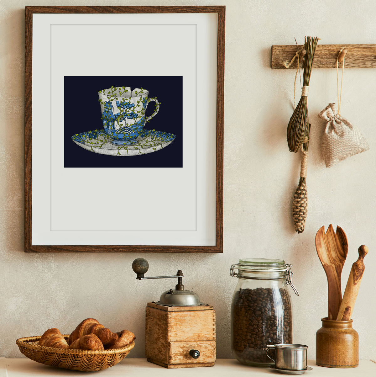 Framed artwork of a teacup on a wall above a kitchen counter with jars, a grinder, and a basket.