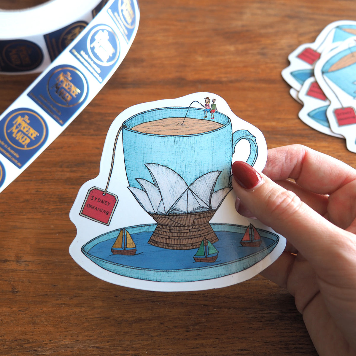 Magnet of a tea cup with a Sydney Opera House design held by a hand on a wooden surface.
