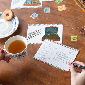 Person writing on a Tasmania postcard with a cup of tea and cookies on a wooden table.