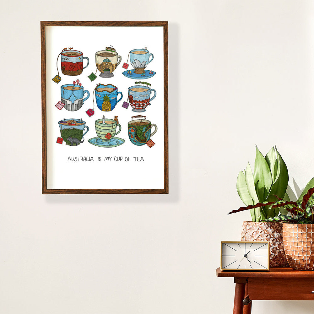 Framed artwork with illustrations of tea cups and text 'Australia is my cup of tea' on a wall above a wooden side table with a clock and plant.