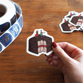 Hand holding a magnet of a lamington with people on it, with a roll of similar magnets in the background.